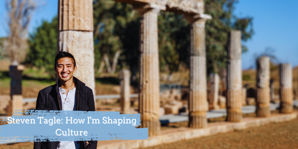 Photograph of a man in his 20s with light skin tone and short black hair. He is standing in front of ancient ruins, columns that are falling down.