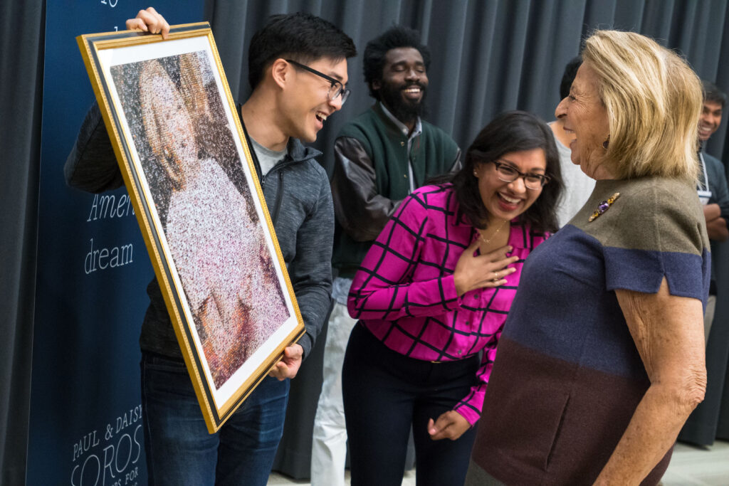 Photograph of a man in his 20s with light skin tone and black hair, holding a portrait that cannot fully be seen, he has an expression of joy on his face. A woman in her 20s with medium skin tone and shoulder length black hair, stands next to him laughing. A man in his 20s with dark skin tone, black hair and beard, stands in the background, also with a big smile.