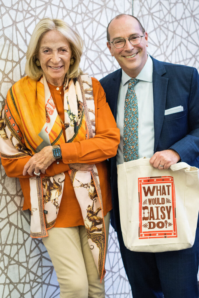 Photograph of two people standing against a wall. On the left a woman in her 80s with light skin tone and shoulder length blonde hair, wearing an orange top with khaki pants and an orange scarf. On the right a man in his 50s with light skin tone and balding hair, he is wearing a navy suit, light green button up and floral tie, he is holding a tote bag that says "What would Daisy do?".