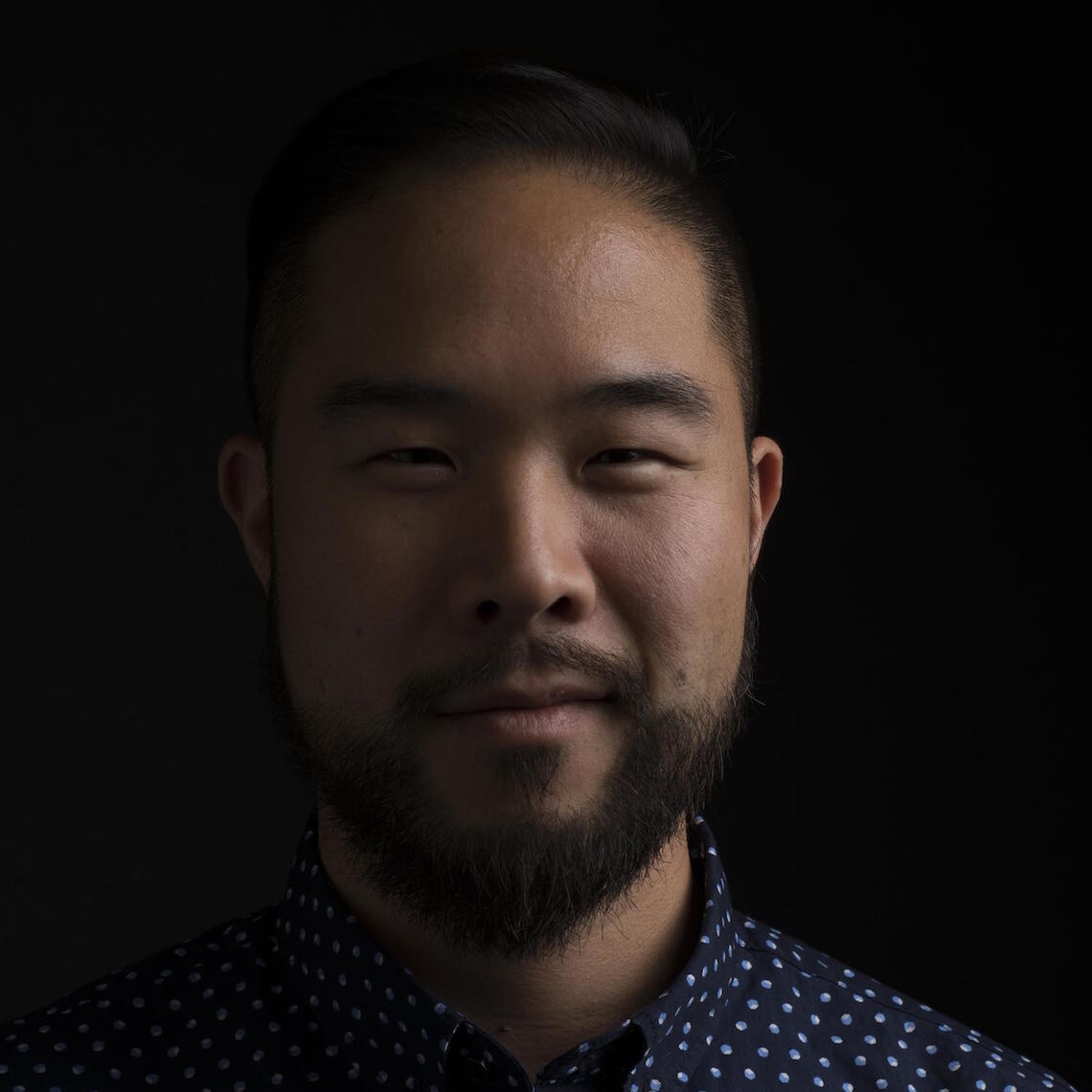 Headshot of a man in his 20s who has heritage from China with light skin tone, black short hair, and a short beard. He is wearing a blue button up shirt and smiling at the camera.