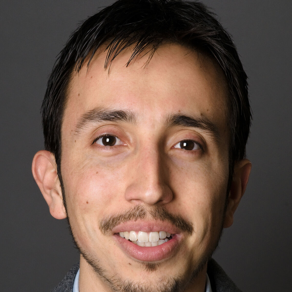 Headshot of a man in his 20s who has heritage from Colombia with light skin tone, short black hair, mustache and goatee. He is smiling at the camera.