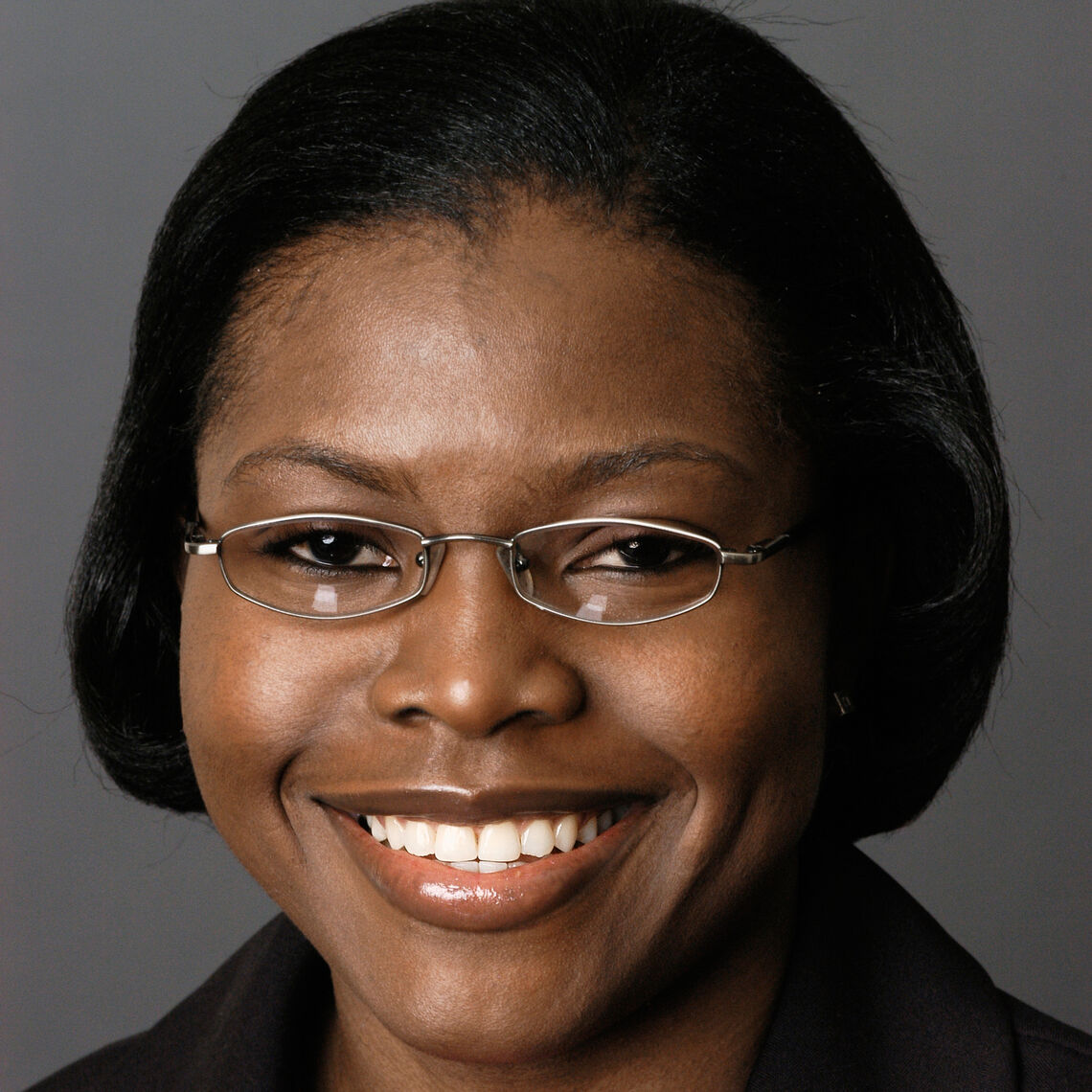 Headshot of a woman in her 20s who has heritage from Nigeria with medium-dark skin tone and black hair cut into a bob. She is wearing a black jacket and small oval silver rimmed glasses. She is smiling at the camera. 