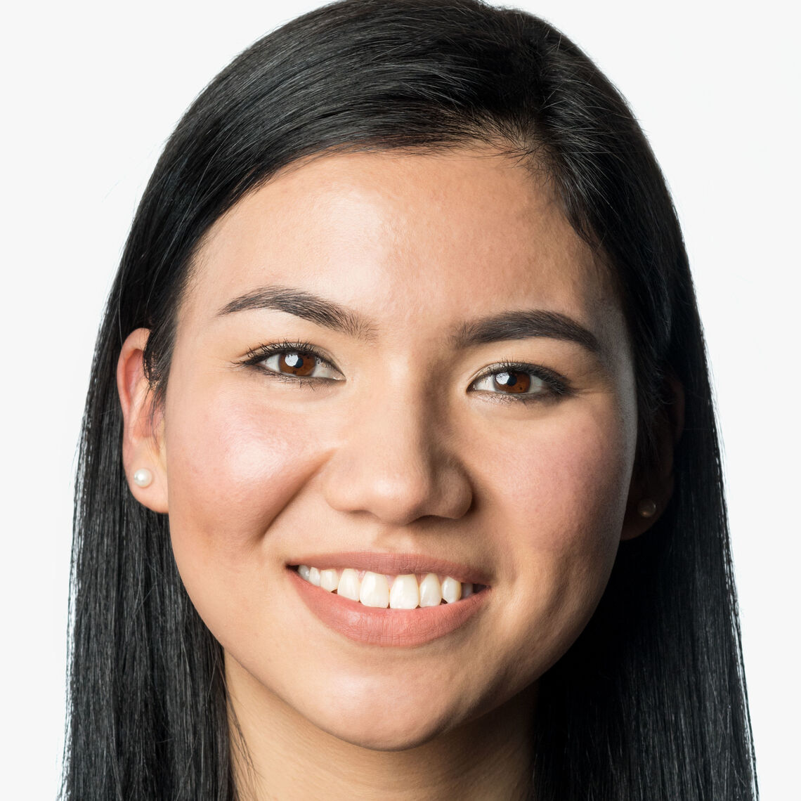 Headshot of a woman in her 20s who has heritage from Pakistan with light skin tone and long black hair parted on the side; she is wearing a black and white patterned high-neck top and pearl stud earrings; she is smiling at the camera