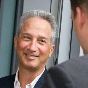 Photo of a man in his 50s with light skin and grey short cut hair. He is wearing a black suit and white button up shirt. He is looking at the person in the foreground and smiling, the camera is looking over their shoulder.