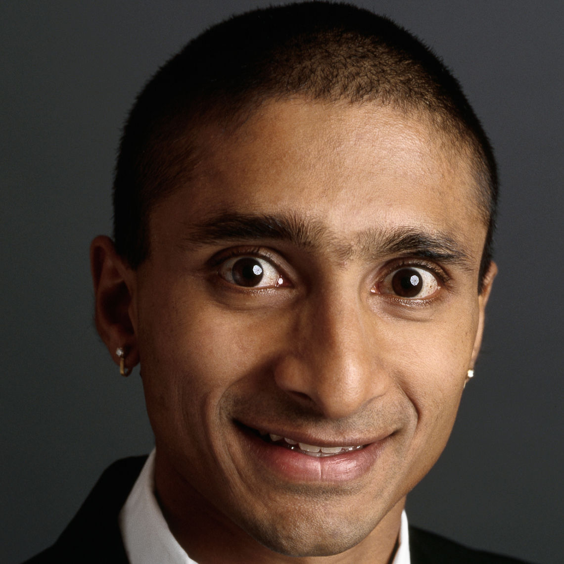 Headshot of a man in his 20s who has heritage from India with medium skin tone and buzzed black hair. He is wearing a black suit, white button up shirt and silver stud earrings. He is smiling at the camera. 