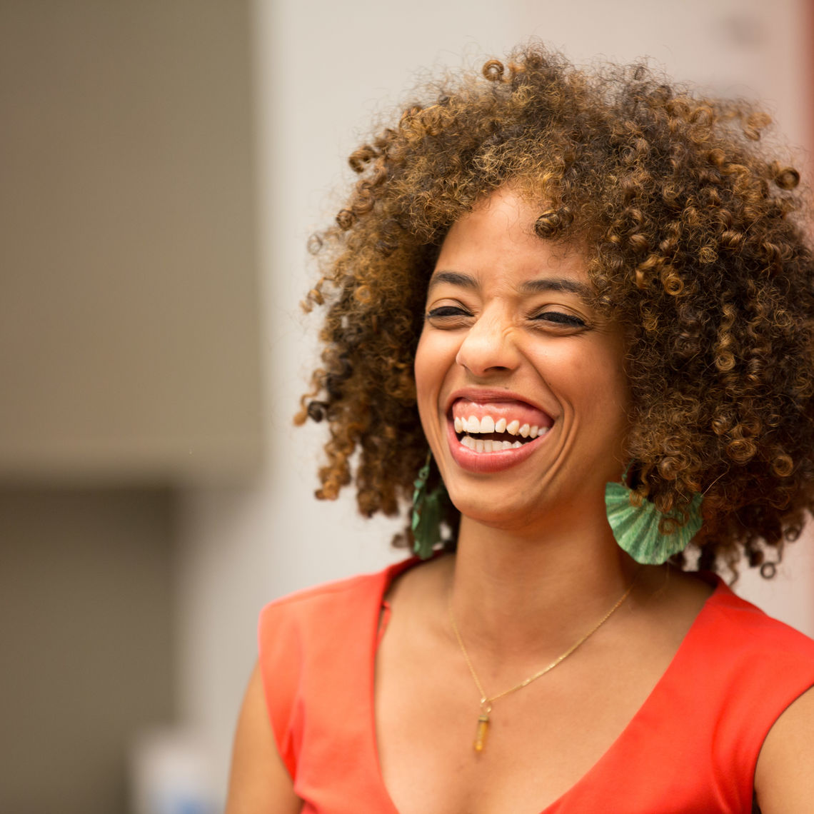 Photograph of a woman in her 30s who has heritage from Antigua and Barbuda and Russia with medium skin tone and curly black and lightly dyed chin length hair. She is wearing an orange top, gold necklace and green copper earrings. She is smiling big and looking off camera. 