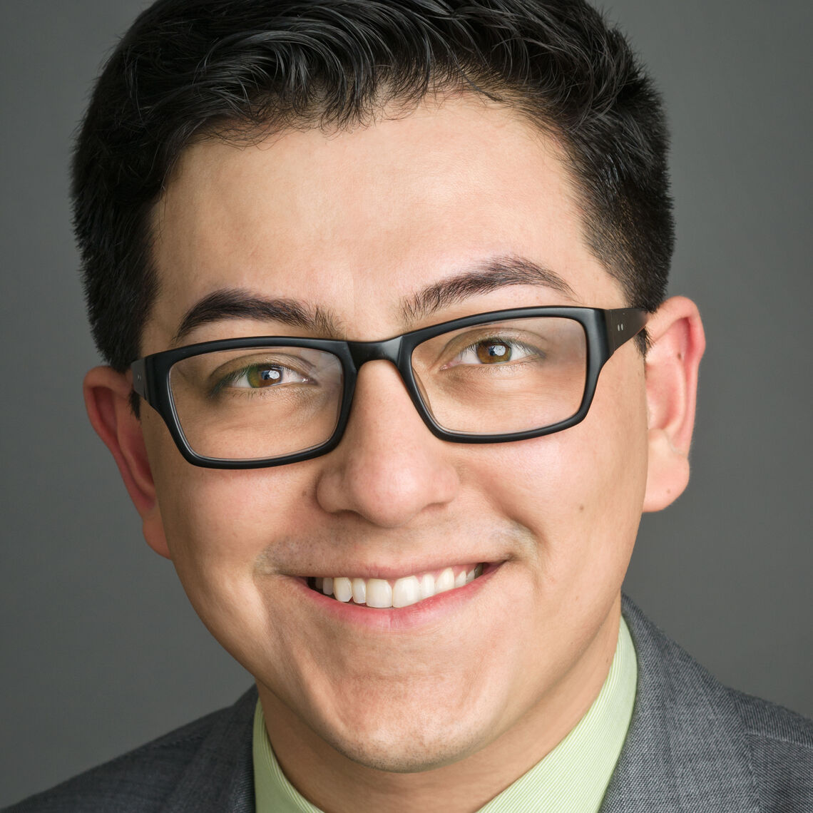 Headshot of a man in his 20s who has heritage from Mexico with light skin tone and black short hair; he is wearing a grey blazer with a light green button up and rectangle medium thickness black glasses; he is smiling at the camera