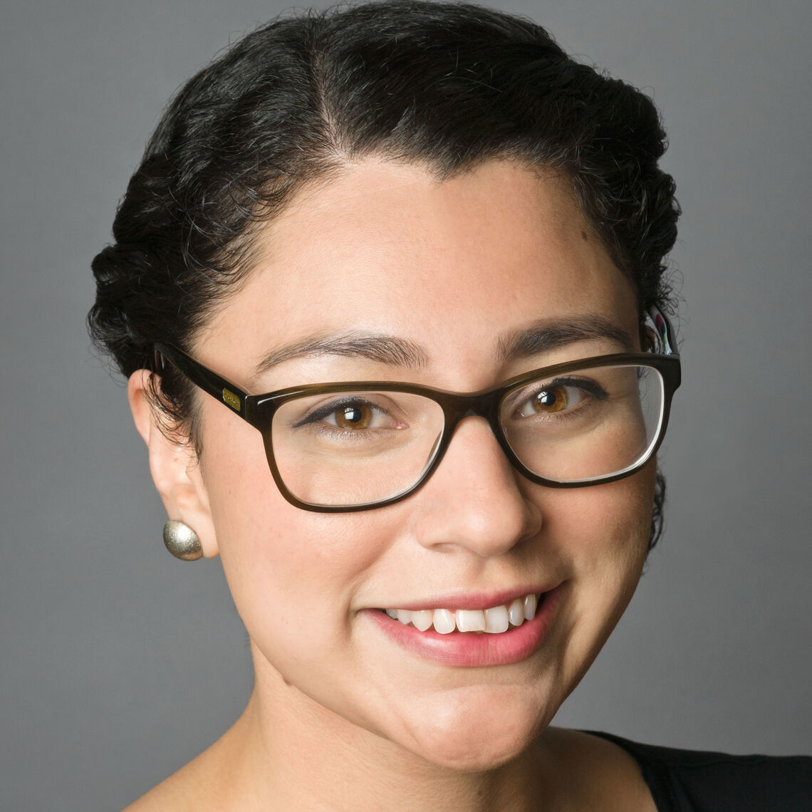 Headshot of a woman in her 20s who has heritage from Honduras and Mexico with light skin tone and black hair, side parted and twisted like a crown and pulled back. She is wearing a black blouse, brass stud earrings and black rimmed glasses. She is smiling at the camera.