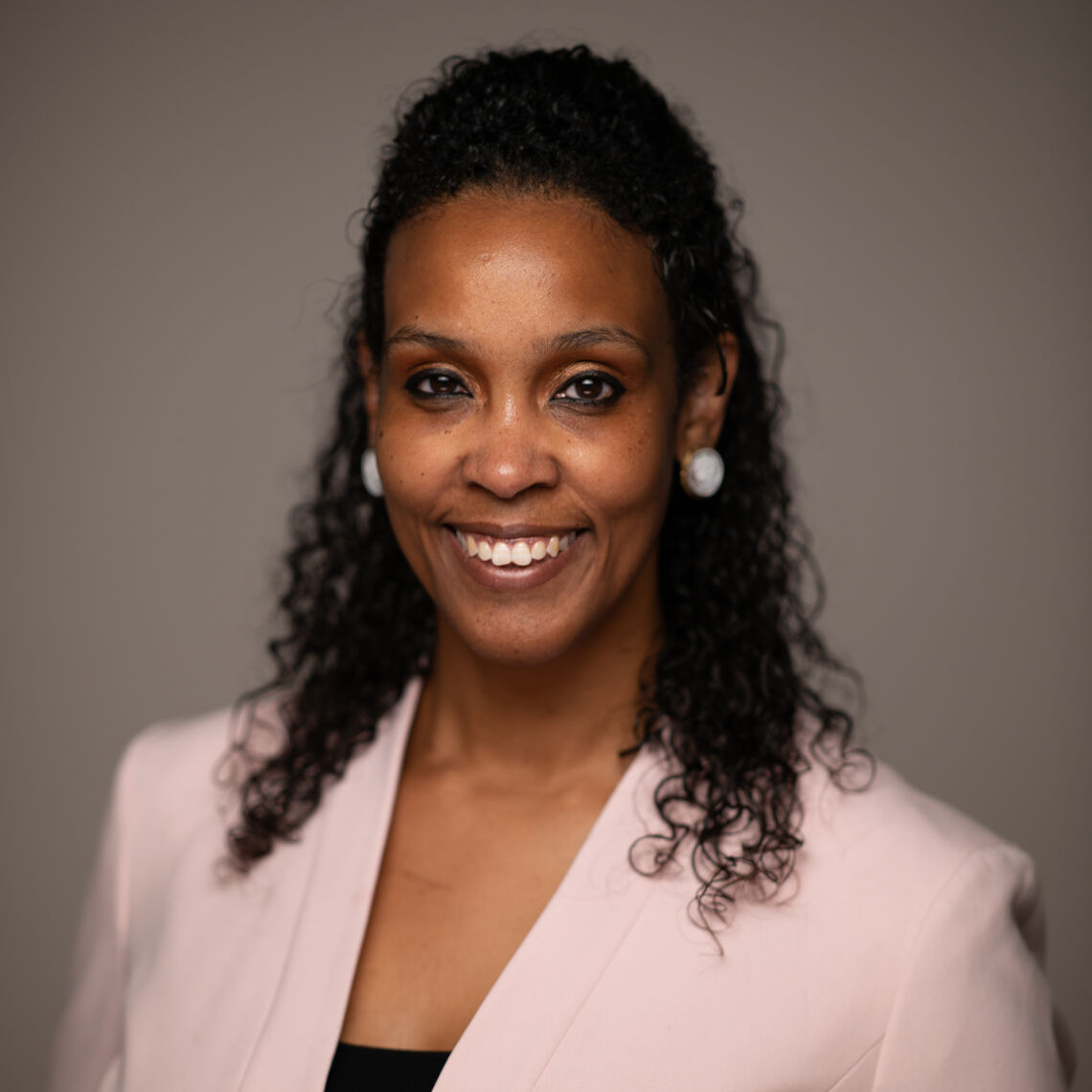 Headshot of a woman in her 30s who has heritage from Jamaica with medium-dark skin tone and shoulder length curly black hair half pulled back. She is wearing a light pink collarless coat with a black shirt underneath and large white earrings. She is smiling at the camera.