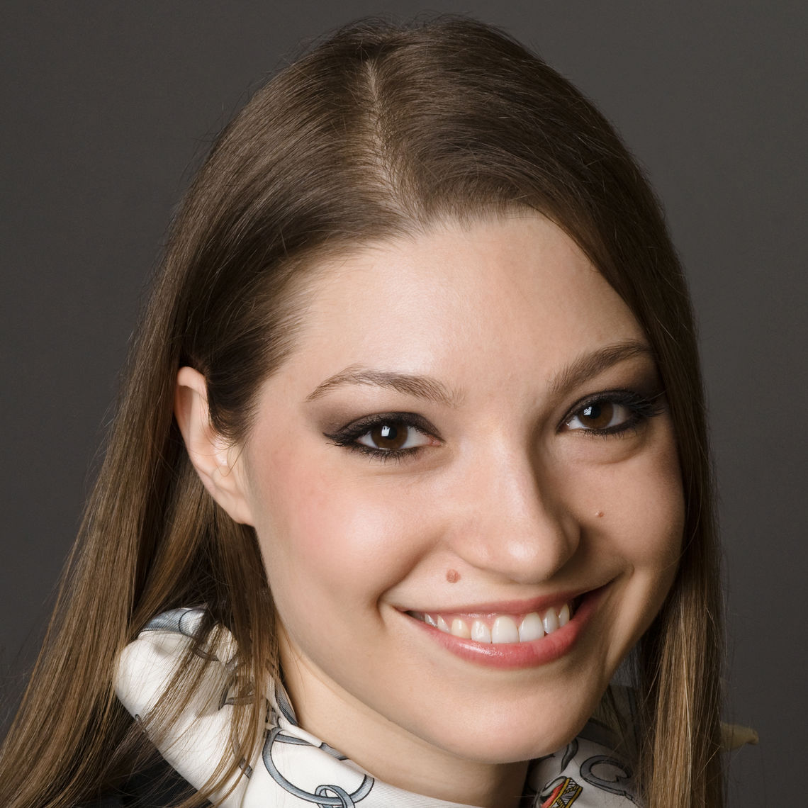 Headshot of a woman in her 20s who has heritage from Croatia with light skin tone and long straight light brown hair. She is wearing a black top and white scarp tied around her neck. She is smiling at the camera. 