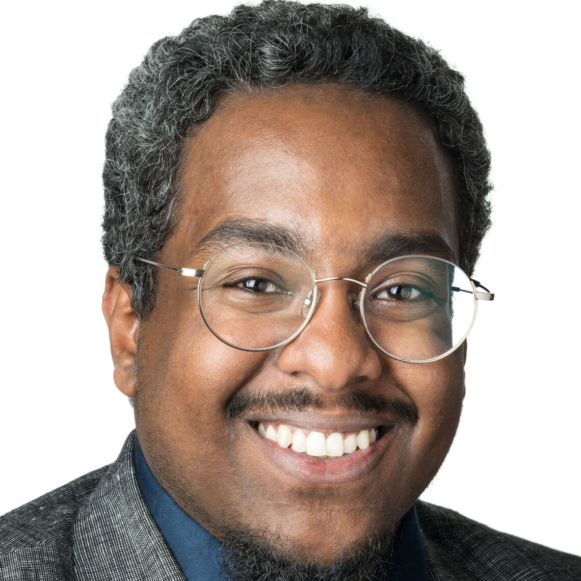 Headshot of a man in his 20s who has heritage from Sudan with medium-dark skin tone, salt and pepper short hair, black mustache and goatee; he is wearing a grey suit, teal button up and white and black tie, he has large round silver thin framed glasses; he is smiling at the camera