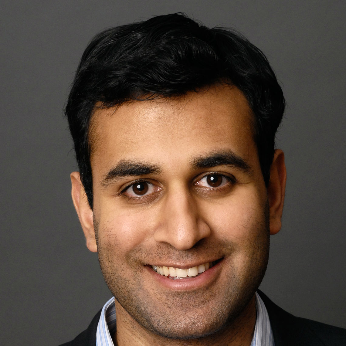 Headshot of a man in his 20s who has heritage from India with medium skin tone and black hair. A black suit and white and blue striped shirt can just be seen. He is smiling at the camera. 