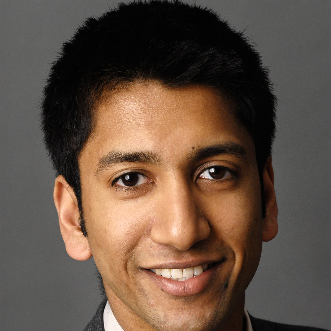 Headshot of a man in his 20s who has heritage from India with medium skin tone and black hair. He is wearing a black jacket and white shirt. He is smiling at the camera. 