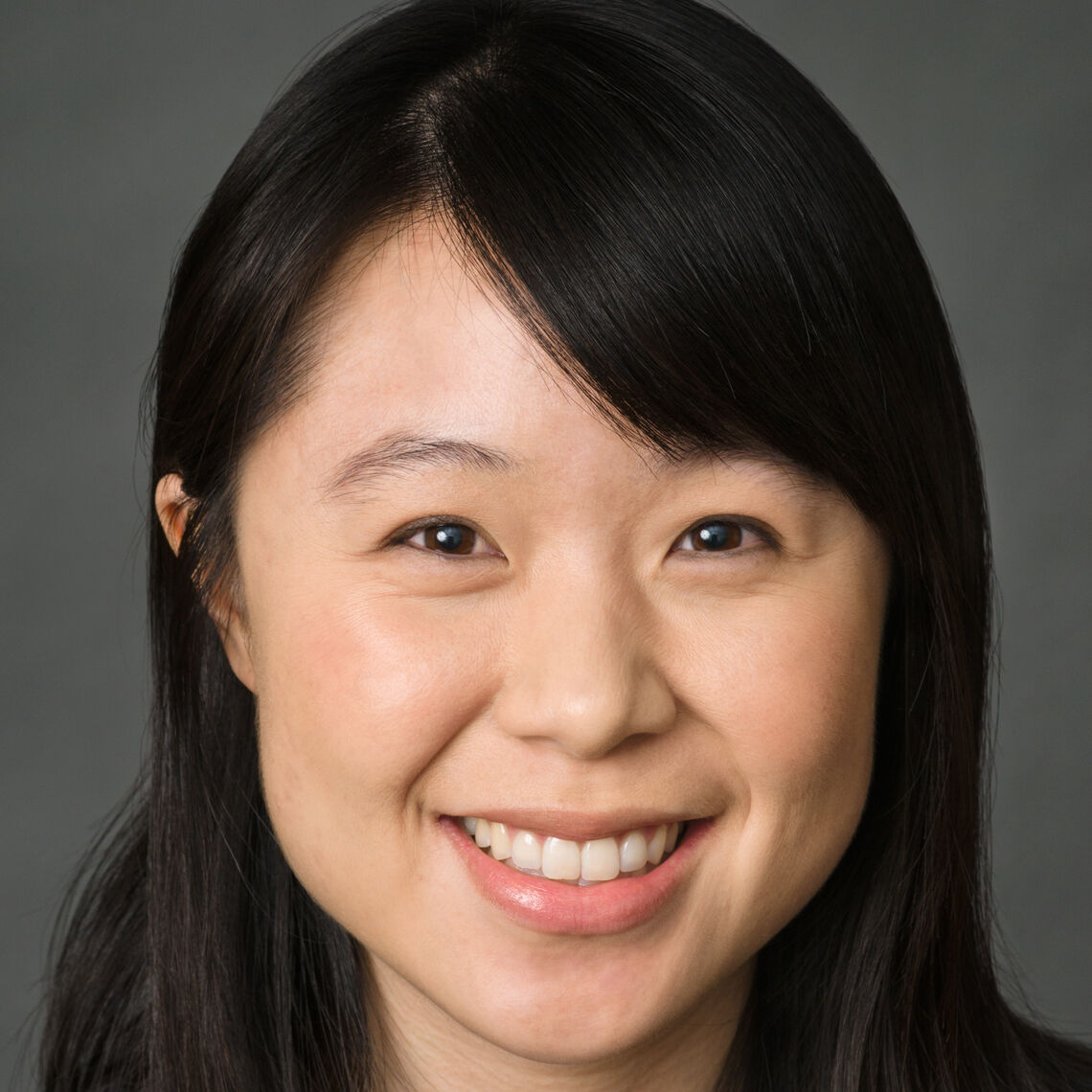 Headshot of a woman in her 20s who has heritage from China with light skin tone and long black straight hair with side bangs, across her left forehead. She is wearing a black top and smiling at the camera. 