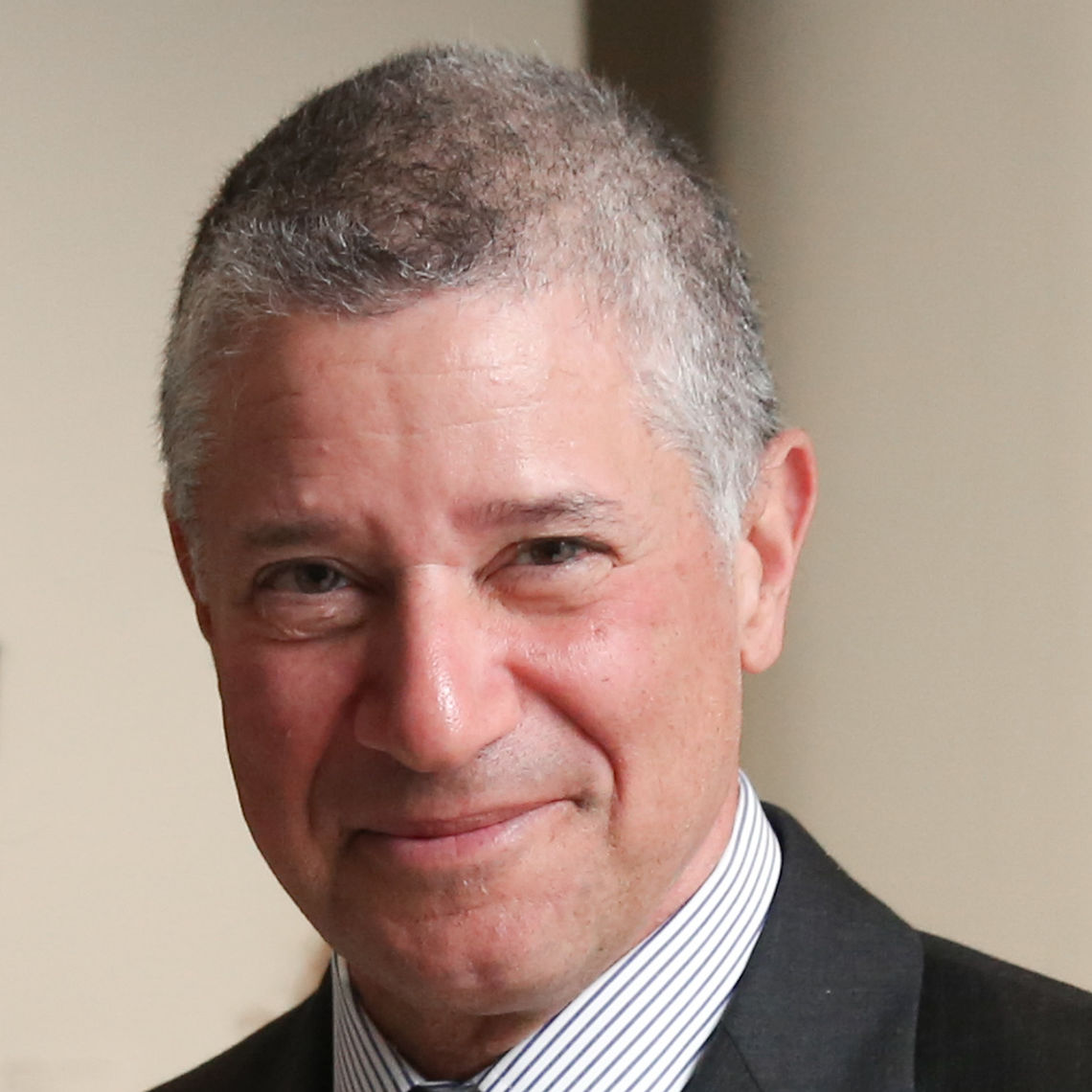 Photo of a man in his 50s with light skin tone and short grey hair. He is wearing a black suit and white striped shirt. He is looking at the camera and smiling. 