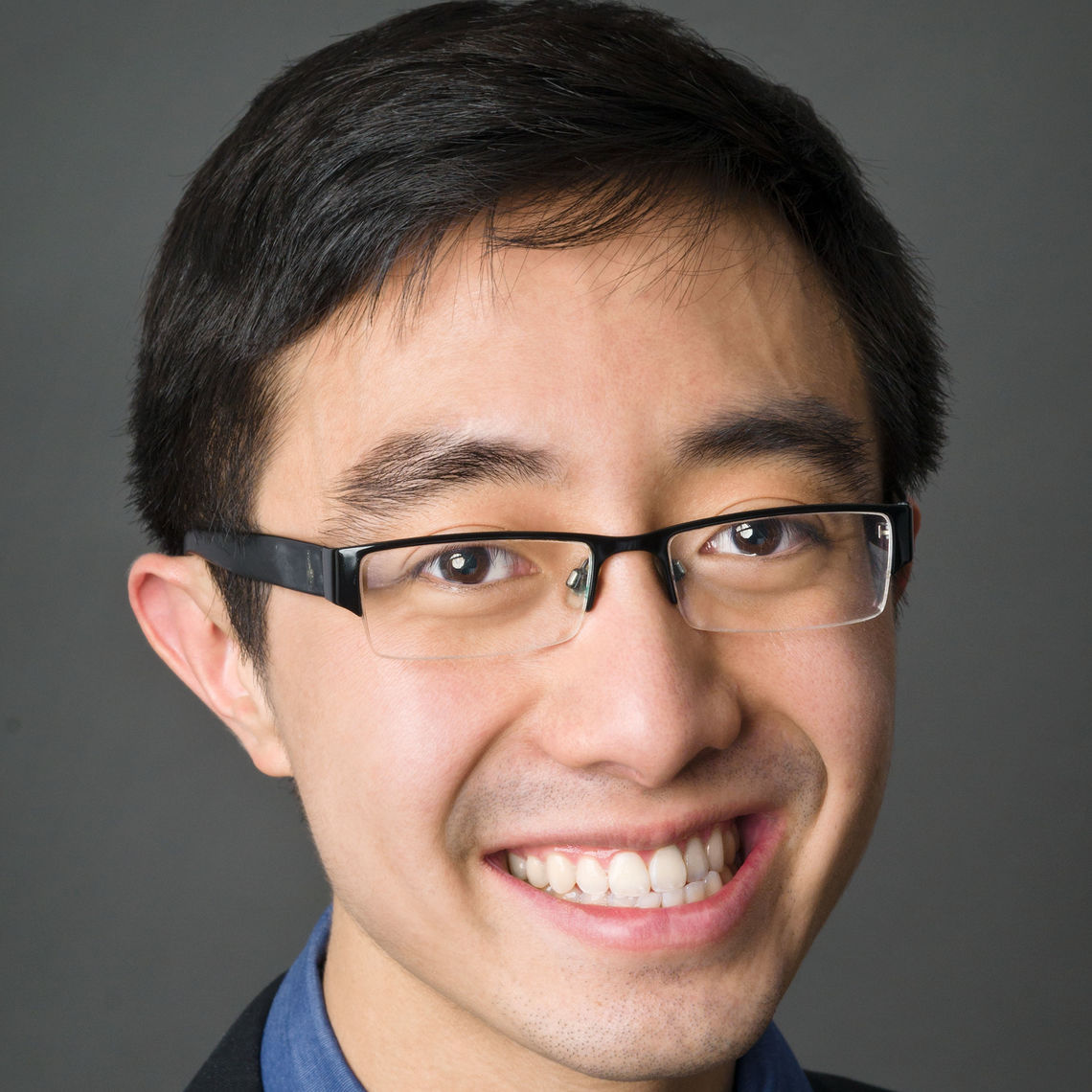 Headshot of a man in his 20s with heritage from Vietnam with light skin tone and black short hair; barely visible in the photo he is wearing a black blazer and blue button up shirt, he wears half-rimmed thin rectangle glasses and smiles at the camera