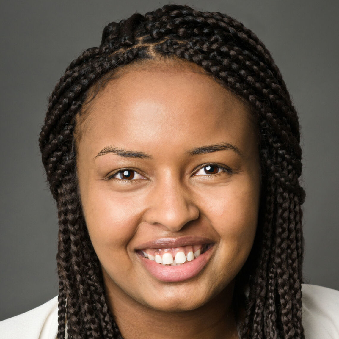 Headshot of a woman in her 20s who has heritage from Somalia with medium skin tone and long black braided hair. She is wearing a white jacket and she is smiling at the camera.