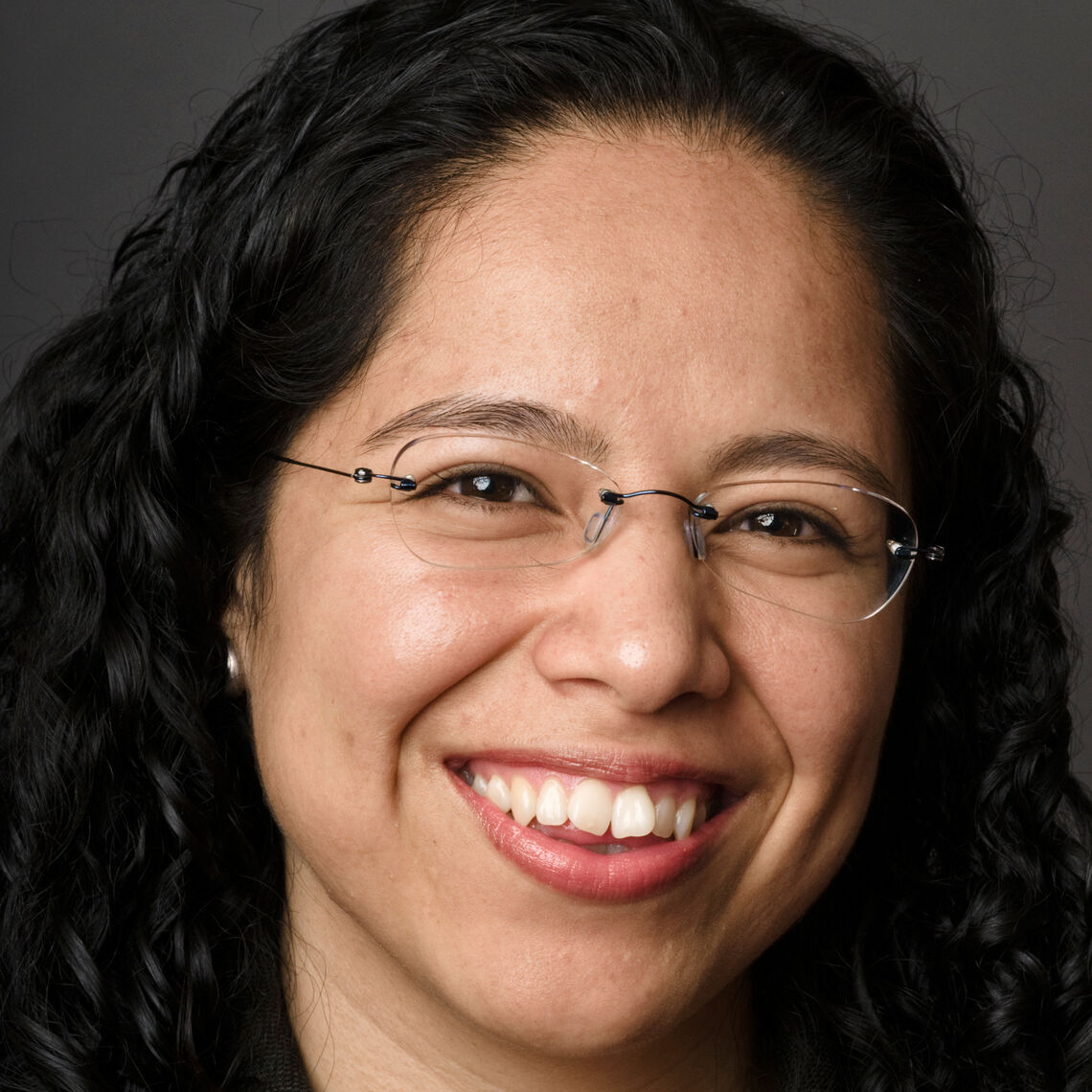 Headshot of a woman in her 30s, who has heritage from Mexico, with light skin tone and long curly black hair. She is wearing no frame rectangle glasses. She is smiling at the camera.