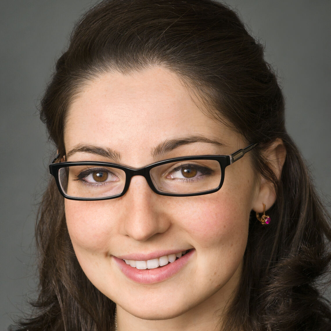 Headshot of a woman in her 20s who has heritage from Azerbaijan with light skin tone and dark brown shoulder length, half pulled back; she is wearing a black top with a sweater that has a black beaded neckline, a silver necklace, red jeweled earrings and thin black rectangle framed glasses; she is smiling at the camera.