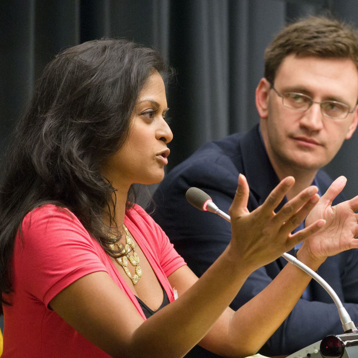 Photograph of a woman in her 30s with heritage from Bangladesh, she has medium skin tone and long black curled hair. She is wearing a bright red t-shirt and gold necklace. She is sitting at a microphone, hands gesturing and looking away from the camera.