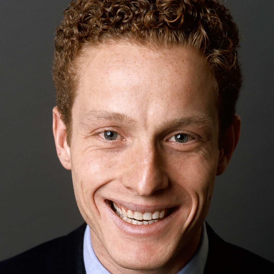 Headshot of a man in his 20s who has heritage from England and Italy who has light skin tone and dark red hair. He is wearing a black suit and smiling at the camera. 