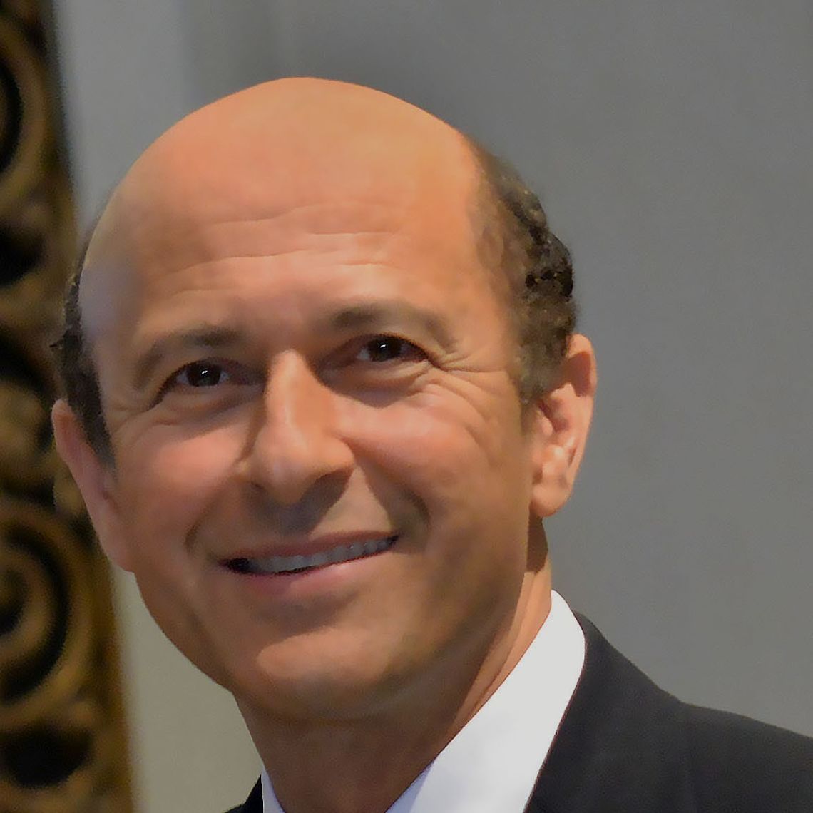 Headshot of a man in his 50s with light skin tone and brown balding hair. He is wearing a black suit and white button up. He is looking at the camera and smiling. 