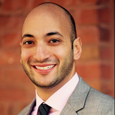 Headshot of a man in his 20s who has heritage from Saudi Arabia with light-medium skin tone, bald and trimmed beard. He is wearing a grey suit and white button up shirt with a tie. He is smiling at the camera. 