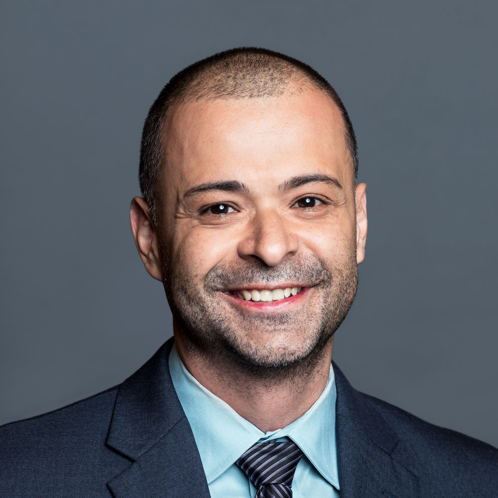 Headshot of a man in his 30s with heritage from Colombia who has light-medium skin tone and buzzed black hair with a trimmed beard. He is wearing a navy suit, light blue button up and tie. He is smiling at the camera. 