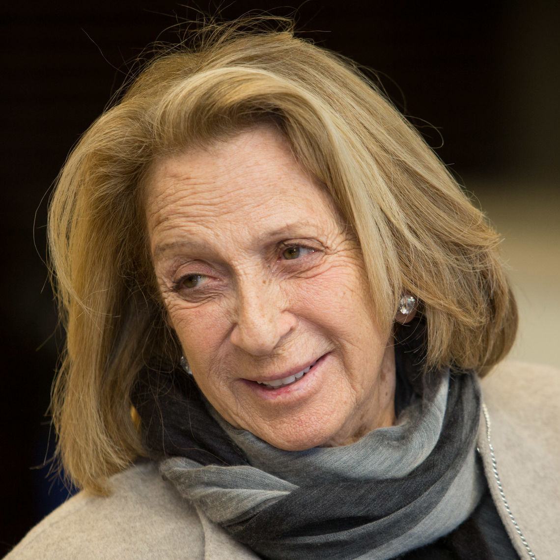 Photo of a woman in her 80s with light skin tone and blonde hair. She is wearing a tan sweater and has a grey scarf wrapped around her neck. She is looking off camera.