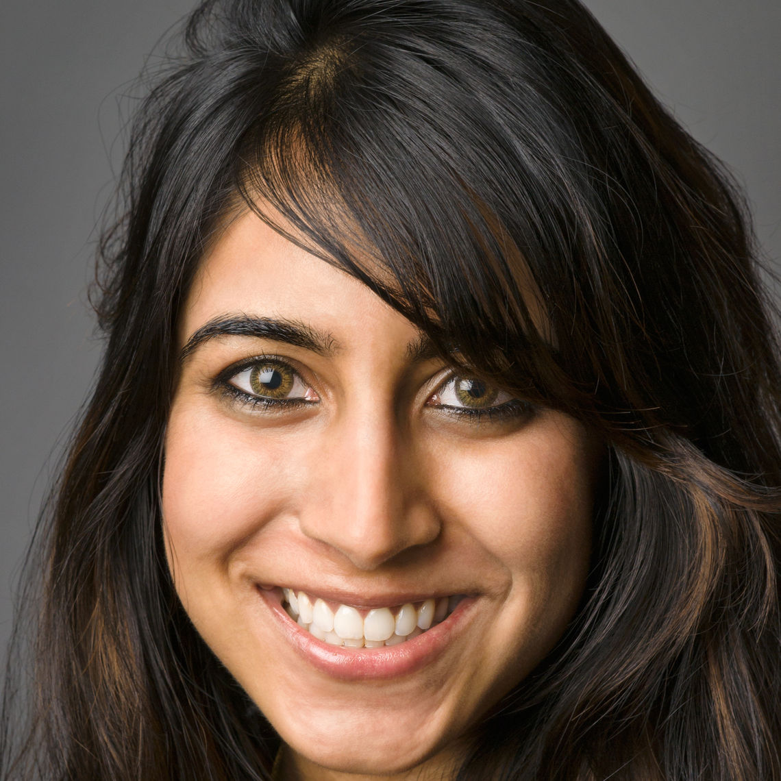 Headshot of a woman in her 20s who has heritage from Tanzania and Uganda with light-medium skin tone and black with dyed red highlights, curled and half pulled back with swoopy bangs. She is smiling at the camera. 