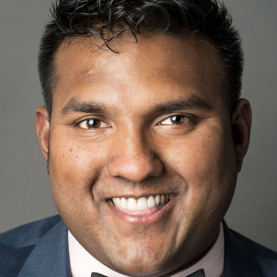 Headshot of a man in his 20s who has heritage from Suriname with medium skin tone and black crew cut hair; he is wearing a blue suit with a white button up and a bow tie.