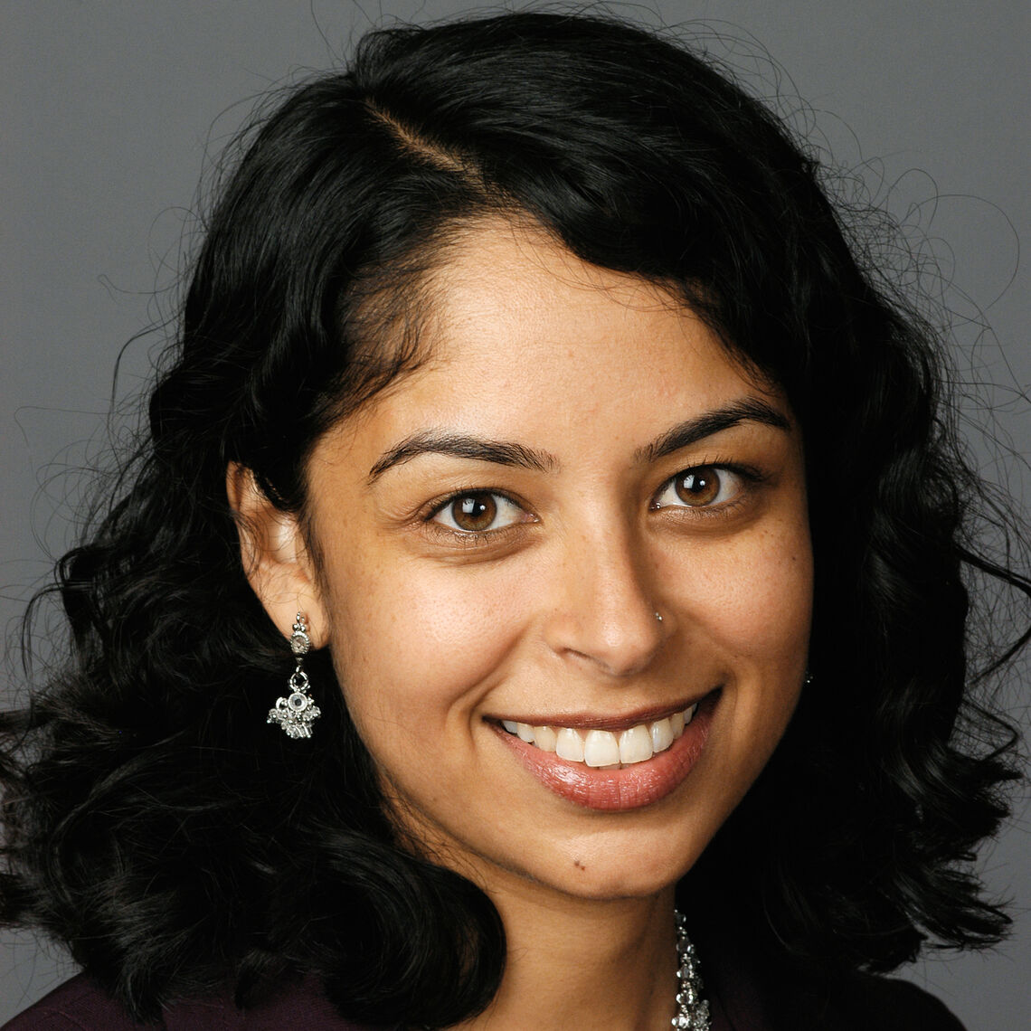 Headshot of a woman in her 20s who has heritage from India with medium skin tone and black shoulder length curly hair with a side part; she is wearing a burgundy blazer with a silver necklace and matching dangly earrings; she is smiling at the camera.