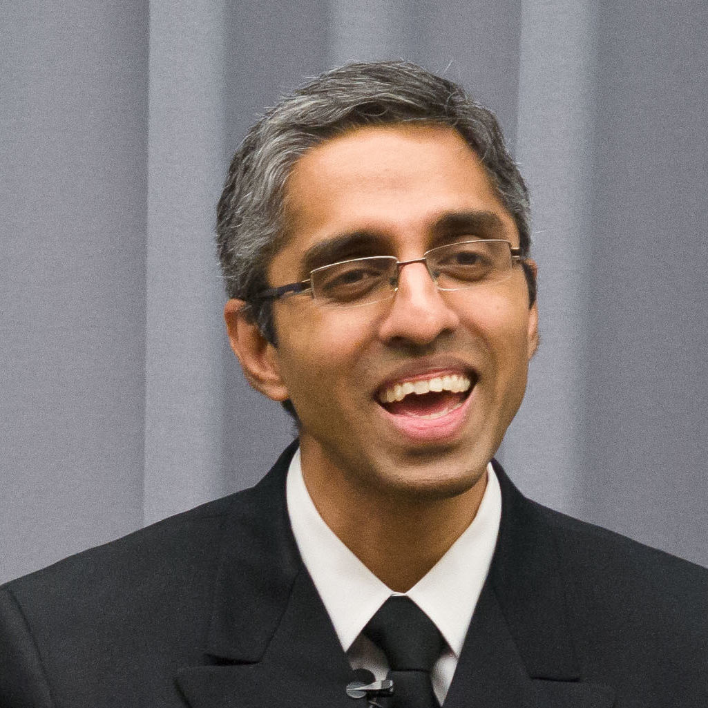 Photograph of a man in his 30s who has heritage from United Kingdom with medium skin tone and salt and pepper hair combed back. He is wearing a navy suit, white button up and black tie.