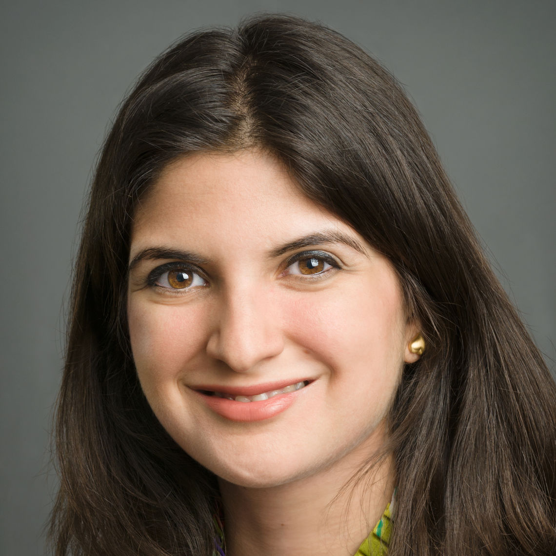 Headshot of a woman in her 20s who her heritage from Iran, with light skin tone and long dark brown hair parted. She is wearing gold stud earrings. She is smiling at the camera. 