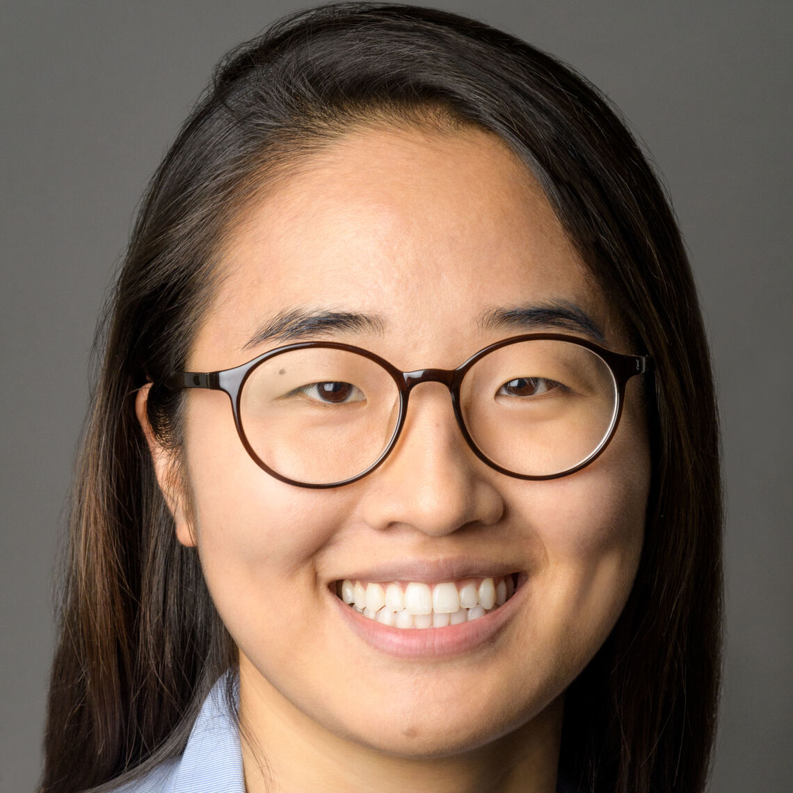 Headshot of a woman in her 20s who has heritage from South Korea, with medium-light skin tone and long straight black hair. She is wearing a light blue button up shirt and round black framed glasses. She is smiling at the camera. 