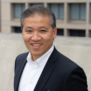 Headshot of a man in his 30s who has heritage from China with light-medium skin tone and salt and pepper combed back hair. He is wearing a black blazer and white button up shirt. He is smiling at the camera.