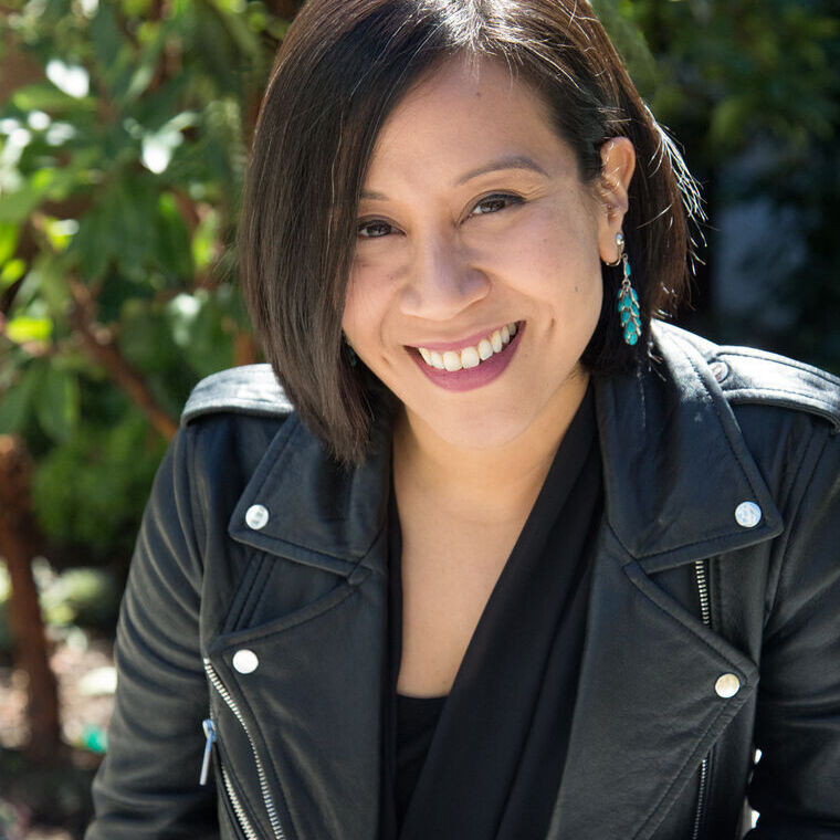 Headshot of a woman in her 30s who has heritage from China and India with light skin tone and chin length dark brown bob. She is wearing a black top, black leather jacket and dangly turquoise earrings. 