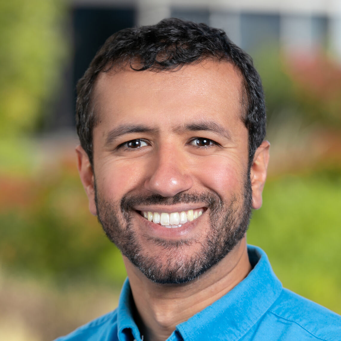 Headshot of a man in his 20s who has heritage from Egypt with light skin tone, black hair and a short beard. He is wearing a blue button up shirt and smiling at the camera. 