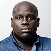 Headshot of a man in his 30s with heritage from Jamaica he has dark skin tone, he is bald. He is wearing a denim button up shirt and he looks directly at the camera.