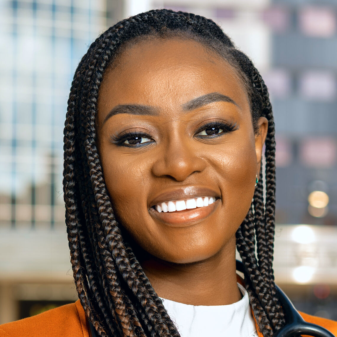 Headshot of a woman in her 20s who has heritage from Nigeria with dark-medium skin tone and long black braided hair. She is wearing a white shirt and orange jacket, smiling at the camera. 