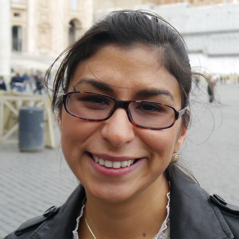 Headshot of a woman in her 30s who has heritage from Mexico with light-medium skin tone and dark brown hair pulled back. She has on a grey jacket and oval tortoiseshell glasses. She is smiling at the camera.