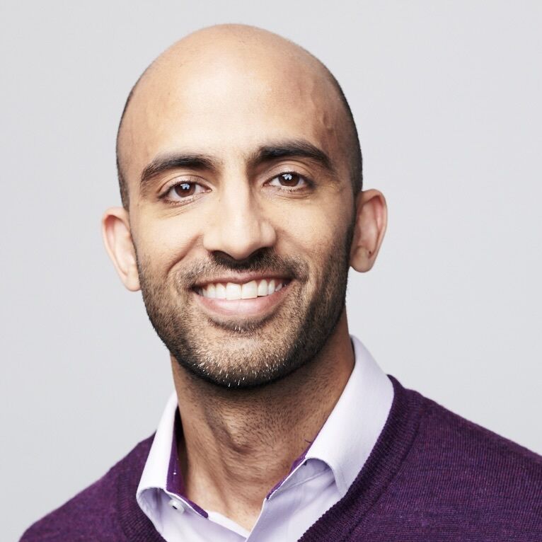 Headshot of a man in his 30s who has heritage from Pakistan, he is bald and has a short beard. He is wearing a light purple button up shirt and a purple v-neck sweater. He is smiling at the camera. 