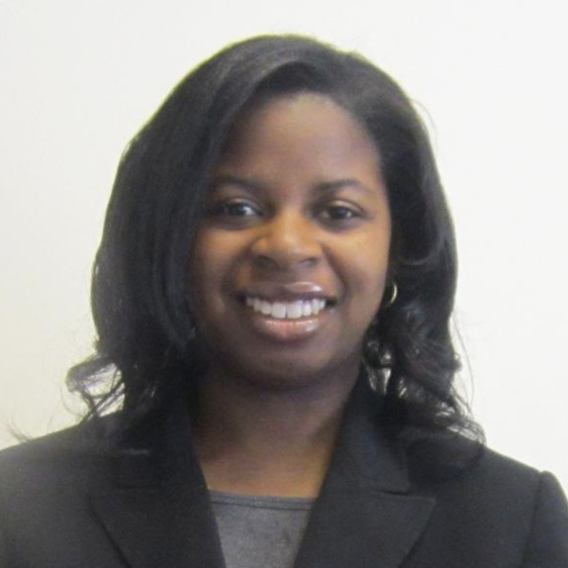 Headshot of a woman in her 20s who has heritage from Nigeria with dark skin tone and black shoulder length curled hair. She is wearing a black blazer and grey shirt, she is smiling at the camera. 