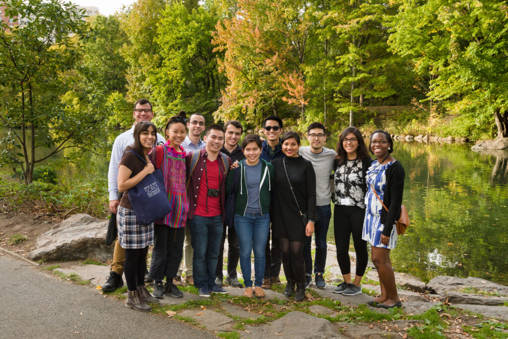 Photograph of twelve people, all in their 20s with varying skin tones and heritages. They are standing in front of a small body of water with trees in the background, the trees are changing color, indicating fall. 