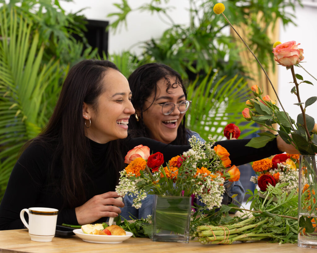 Two women in their 20s sit side by side laughing not looking at the camera, they are surrounded by orange and red flowers with lots of greenery behind them. 