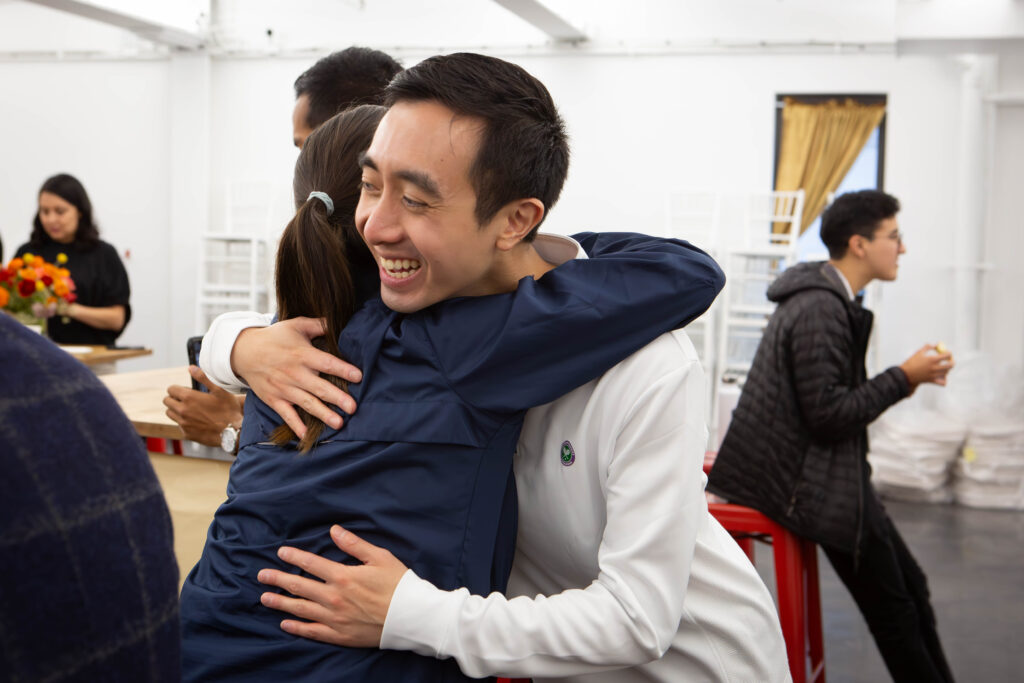 A man and woman hug, her back is to the camera and he is looking past the camera. There are other people standing around them.