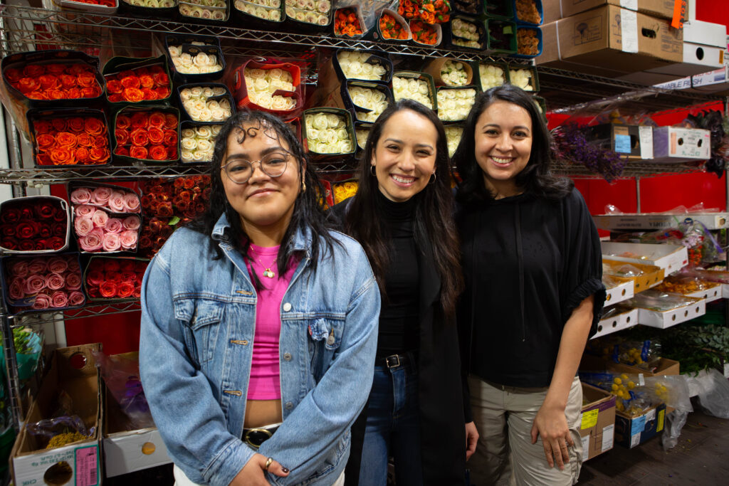 Three women in their 20s stand in front of a wall of packed flowers, smiling at the camera. 