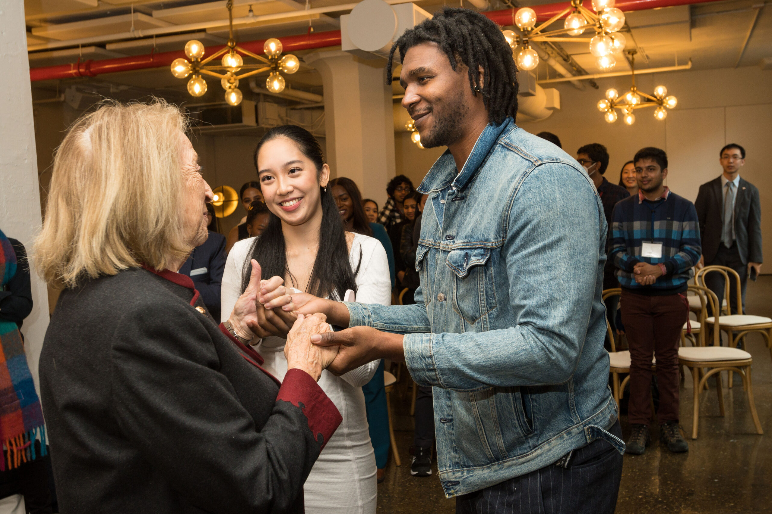 Photograph of a woman in her 80s with light skin tone and blonde shoulder length hair, wearing a black with red accent coat, looks away from the camera and is holding hands with a man in his 20s with medium-dark skin tone and chin length black hair, wearing a jean jacket. They look at each other with admiration. A woman in her 20s with light skin tone and long black hair stands by watching and smiling. 