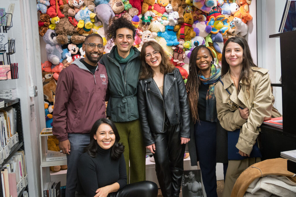 Six people stand posing and smiling in front of a wall of stuffed animals - they are a diverse group of men/women as well as a variety of heritages. 