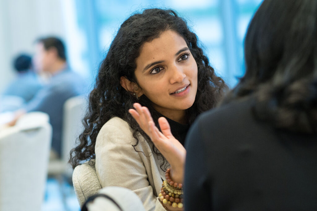 Photograph of a woman in her 20s with medium skin tone and long black curly hair, engaged in conversation while seated. We can see the back of another person in the foreground.